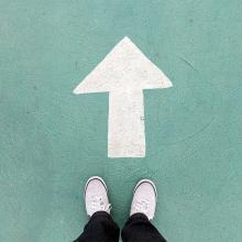 photograph of person's shoes as they satnd in front of upward arrow painted on turquoise colored background