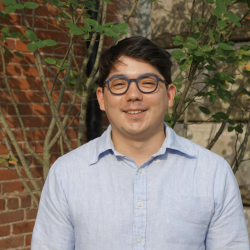 Ian smiles, wearing round-rimmed glasses and a light blue shirt